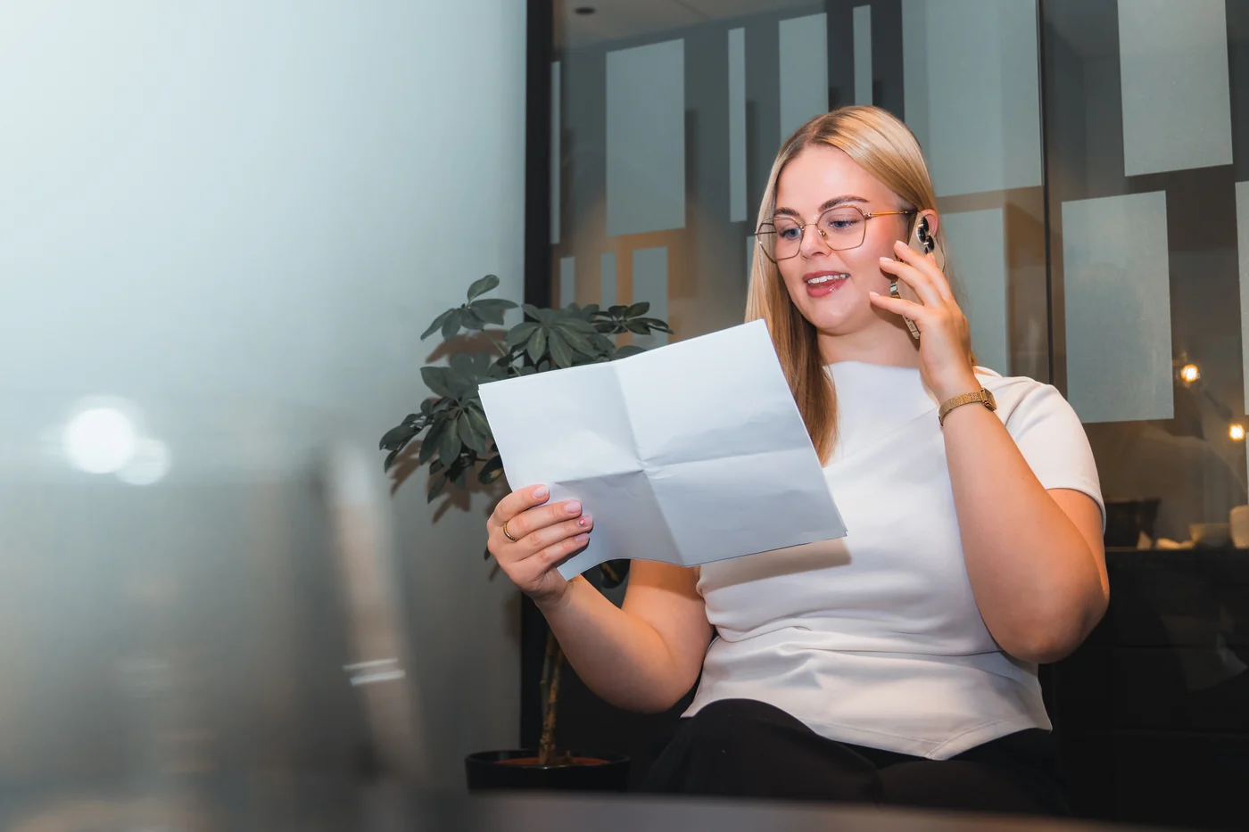 Kvinne med briller sitter i et kontorlandskap, snakker i telefonen og leser et brev om renten på lånet sitt